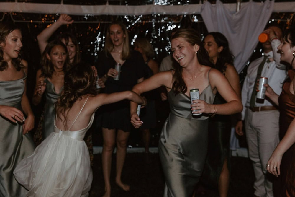 A group of people in formal attire dancing and holding drinks at a celebration, with two women in the foreground holding hands and smiling.
