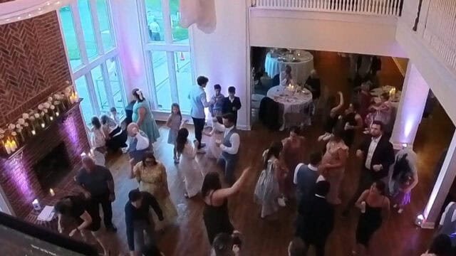 A group of people dance in a decorated hall with draped fabric on the ceiling, large windows, and a DJ booth visible in the foreground.