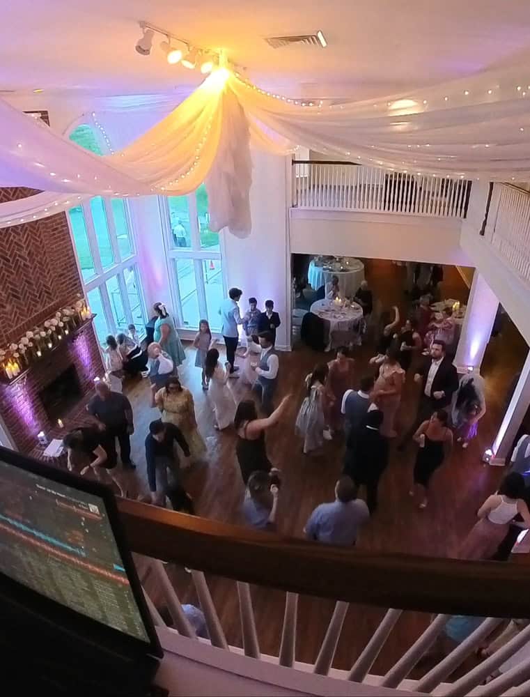 A group of people dance in a decorated hall with draped fabric on the ceiling, large windows, and a DJ booth visible in the foreground.