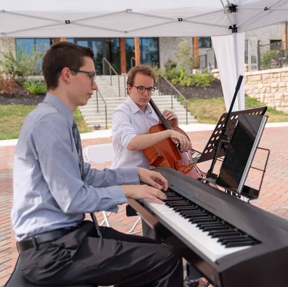 Two musicians perform outdoors under a canopy; one plays an electric keyboard while the other plays a cello, with sheet music in front of them.