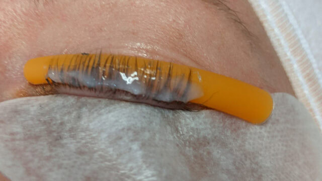 Close-up of a woman's closed eye with a yellow silicone rod and perming solution applied to the eyelashes, indicating a lash lift procedure in progress.