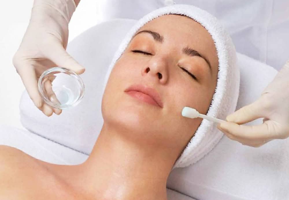 Woman receiving a facial treatment; eyes closed, wearing a towel headband, while a gloved practitioner applies a liquid with a cotton swab.