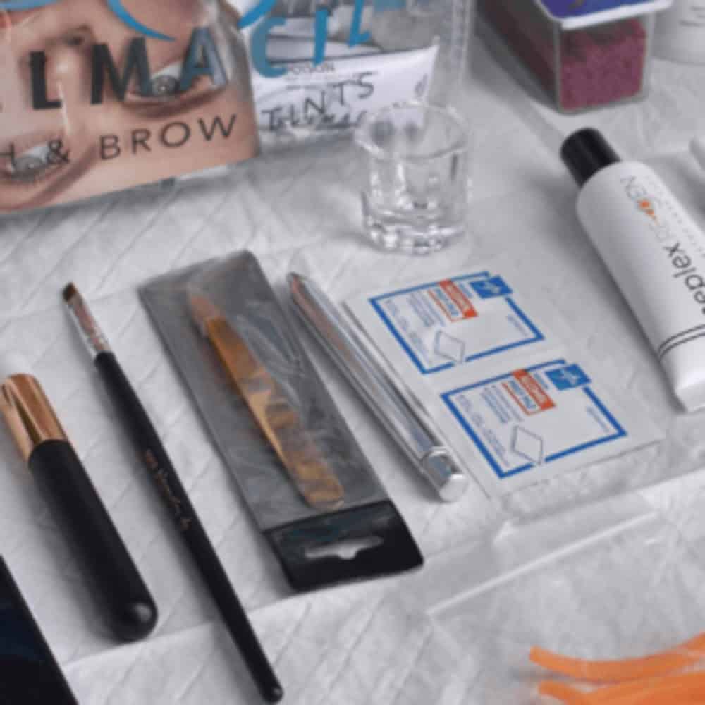 A collection of beauty tools and supplies, including makeup brushes, brow tint packaging, cleansing pads, and a small clear glass, arranged on a white surface.