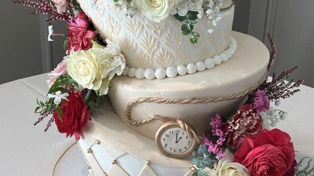 Three-tiered white cake with gold accents, decorated with red, pink, and white flowers, and a small pocket watch inserted on the middle tier.
