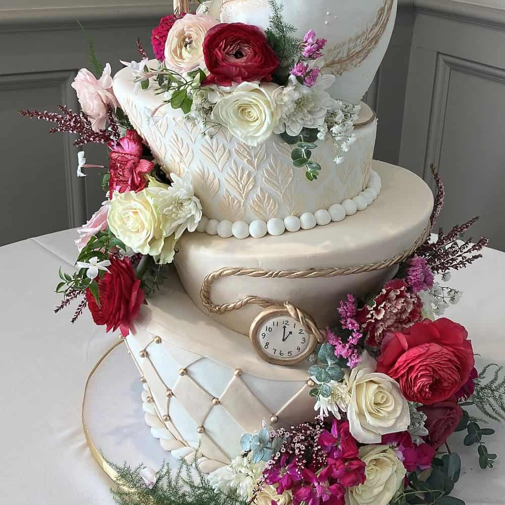 Three-tiered white cake with gold accents, decorated with red, pink, and white flowers, and a small pocket watch inserted on the middle tier.