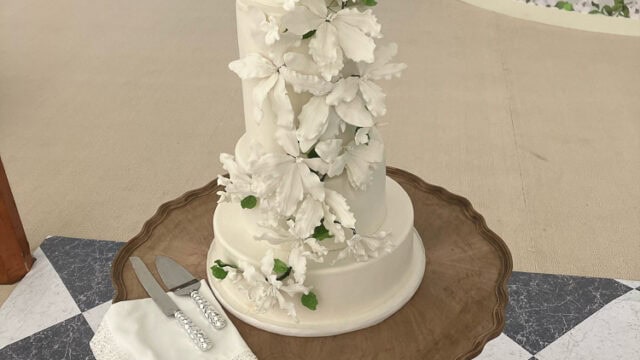 A four-tiered white wedding cake decorated with cascading white flowers and green leaves sits on a wooden table with a cake knife and server on a napkin.