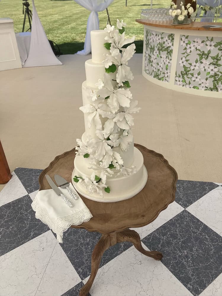 A four-tiered white wedding cake decorated with cascading white flowers and green leaves sits on a wooden table with a cake knife and server on a napkin.