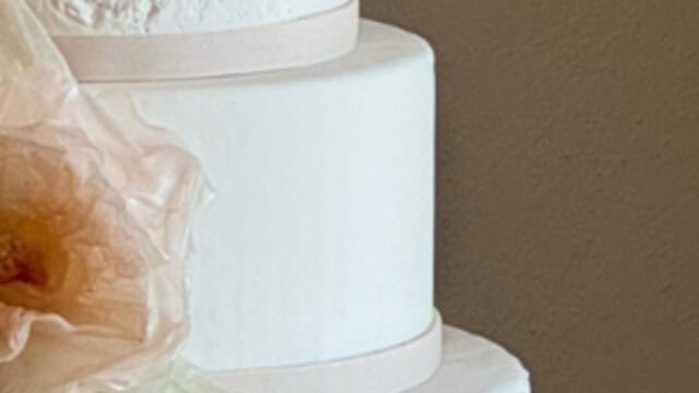 Three-tiered white wedding cake with smooth icing and minimal decoration, featuring a large, delicate, pale flower accent on the side.