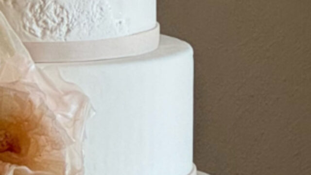 Three-tiered white cake with smooth icing, accented by thin, light ribbons and a sheer, peach-colored decorative element on the left side.