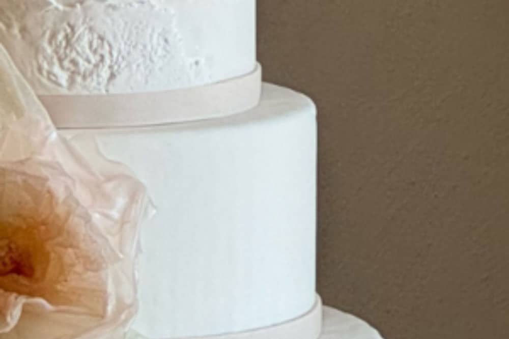 Three-tiered white cake with smooth icing, accented by thin, light ribbons and a sheer, peach-colored decorative element on the left side.