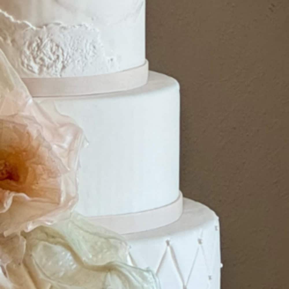 Three-tiered white wedding cake with smooth icing and minimal decoration, featuring a large, delicate, pale flower accent on the side.
