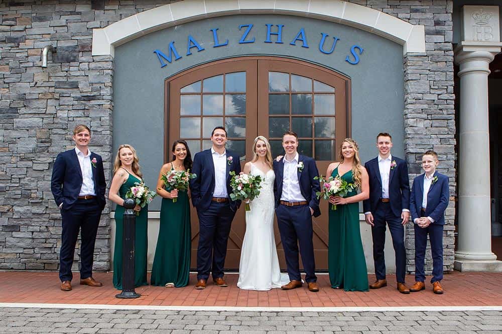 A wedding party of eight people stands in formal attire in front of a stone building with "MALZHAUS" written above double doors.