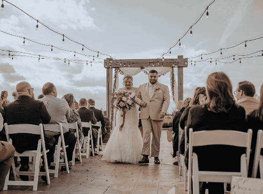 A bride and groom walk down the aisle together outdoors, surrounded by seated guests under string lights, with a wooden arch in the background.