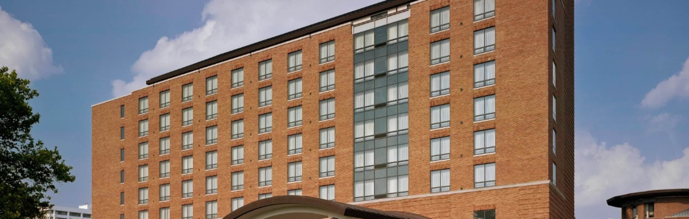 A multi-story red brick hotel building with many windows and a curved entrance canopy, set against a blue sky with scattered clouds.